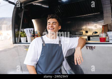 Cheerful food truck owner leaning counter. Young guy working as a chef in a food truck standing with closed eyes and smiling. Stock Photo