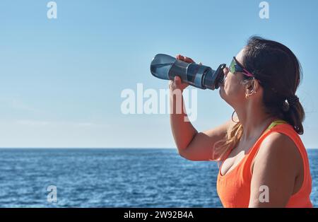 Latin woman, middle-aged, resting, regaining strength, eating, drinking water, after a gym session, wearing orange top, burning calories, keeping Stock Photo