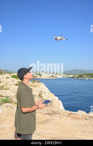 Happy teen boy, flying drone on mediterranean coast, against blue sky during sunny day Spain, Balearic Islands Stock Photo