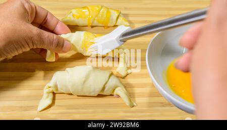 Preparation of croissants, stuffed with ham and cheese and painted with egg yolk, on a wooden board, ready to bake Stock Photo