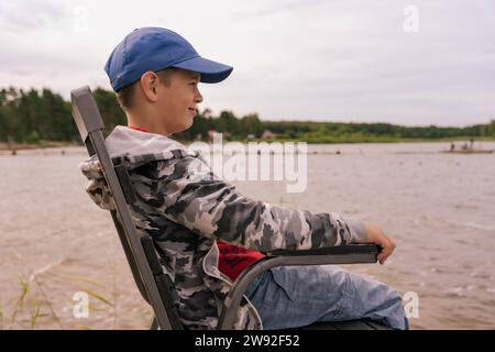 A school-age boy in solitude sits on a chair at the calm lake shore, reflecting on the serene beauty of nature, surrounded by peaceful tranquility and Stock Photo