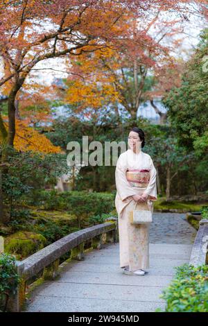 A woman wearing kimono standing on a Japanese style stone arch bridge. Kyoto, Japan. Maple leaves turning red in the autumn season. Fall foliage. Stock Photo