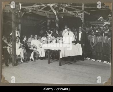 Festive gathering with Dutch and Surinamese, 1925 - 1927 photograph Festive gathering with Dutch and Surinamese. Three men standing at a table, left in the front row Edith Jetten in a chair, on the right a few scouts. Photo with corners attached to a page of an album magazine in the photo album about the Jetten family and the life and working in the Mensstad Moengo in Suriname around 1927. Suriname photographic support  anonymous historical persons portrayed in a group, in a group-portrait Suriname Stock Photo
