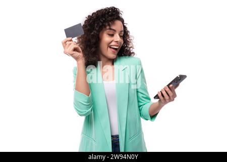 young brunette curly business woman in a jacket uses internet banking and a card in a smartphone Stock Photo