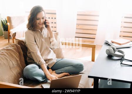 Multitasking in Style. Busy Young Woman on Sofa with Smartphone and Laptop. Sitting on the sofa in her living room, making calls on her smartphone and Stock Photo