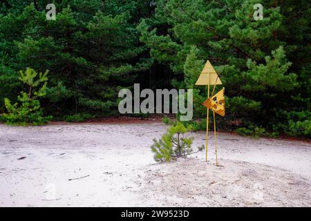 A yellow and black warning sign in a forest, trees. The sign is attached to a post and has trees in the background. Radiation pollution sign near tree Stock Photo