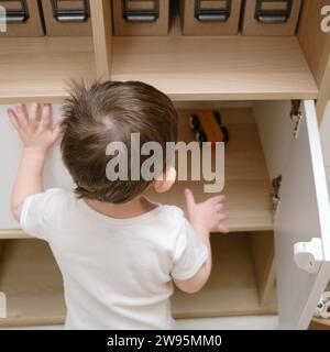 Toddler baby opens the closet door in the home living room. A small child opens a shelf drawer. Kid aged one year eight months Stock Photo