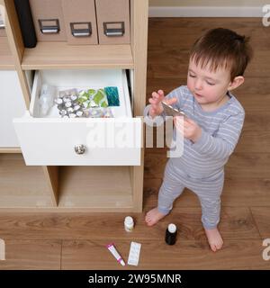 Toddler baby opened the cabinet drawer with pills and medicine. Child boy holding a pack of pills in the home living room. Kid age one year nine month Stock Photo
