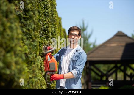 Professional gardener trimming, shaping evergreen thuja hedge with modern electric trimmer at summer time. Portrait of smiling worker looking at camera, while pruning. Concept of gardening services. Stock Photo