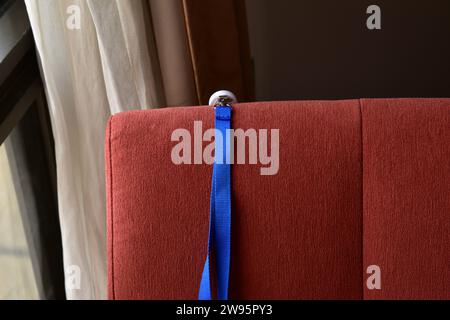 Close-up of a blue keycord hanging over the backrest of red fabric chair with a window and white curtains in the background Stock Photo