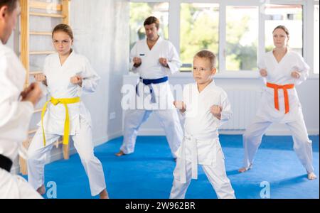 Boy with his family in kimonos and colored belts practicing karate with punches during group martial arts class in gym, accompanied by trainer Stock Photo