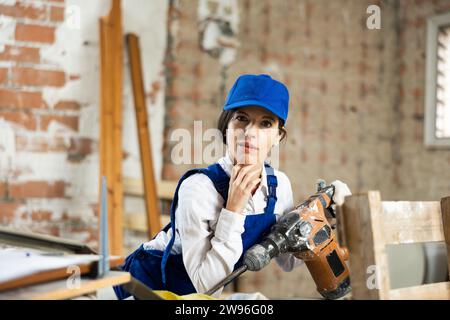 Focused female contractor wearing blue workwear standing with handheld demolition hammer inside building under construction Stock Photo