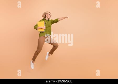 Full length photo of optimistic woman dressed green shirt brown pants flying with book achieving goals isolated on beige color background Stock Photo
