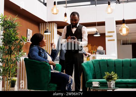 Young African American woman tourist sitting at luxury hotel lobby making order talking with waiter, ordering food and drinks while waiting for check-in. Customer service in hospitality Stock Photo