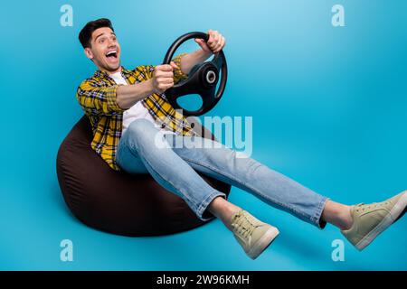 Full body photo of enjoying speed young guy driving automobile simulator using steering wheel in bean bag isolated on blue color background Stock Photo