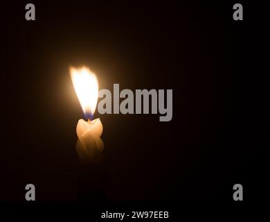 A single burning candle flame or light is glowing on a spiral white candle against little blowing wind on black or dark background on table in church Stock Photo