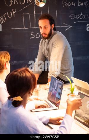 The image captures a candid moment during a start-up meeting, with a focus on a man leaning forward, engaging with his colleagues. In the foreground, a woman is seen typing on a laptop, her attention directed towards the screen where another team member is present via video call. The backdrop is a chalkboard with words and diagrams scribbled on it, suggesting brainstorming and strategic planning. This scene reflects the collaborative spirit of a modern work environment. Collaborative Efforts, Start-up Meeting in Progress. High quality photo Stock Photo