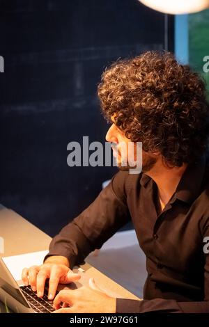 The image presents a profile view of a focused individual working intently on a laptop. His curly hair and the sharp attire suggest a creative professional possibly immersed in software development or digital content creation. The ambient lighting highlights his concentration and the seriousness of his work. The dark backdrop provides a contrast that puts emphasis on the subject and his task. Deep in Development, Software Engineer at Work. High quality photo Stock Photo