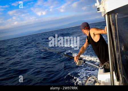 The young man in a baseball cap onboard the vessel in the course of fishing against the background of the blue sea. Stock Photo
