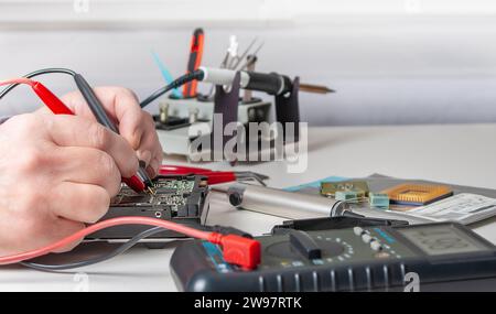 Close-up of a technician's hands in a workshop. The repairer is using a multimeter to measure the parameters of an electronic device on a table. Stock Photo