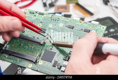 Close up of a technician's hands in a workshop. Repairer is soldering circuit board of electronic device on table with tweezers. Stock Photo