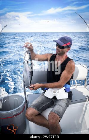 The young man in a baseball cap onboard the vessel in the course of fishing against the background of the blue sea. Stock Photo