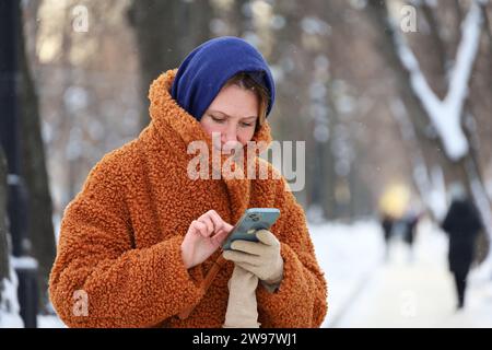 Woman in warm clothes standing with smartphone in hands on winter city street during snow Stock Photo