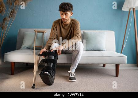 A young man with an ankle injury wearing a rigid ankle orthosis sits on the sofa with crutches at home Stock Photo
