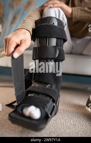 Young Man attaching an ankle orthosis to his leg to support ligaments while sitting on the sofa at home. Medicine concept Stock Photo