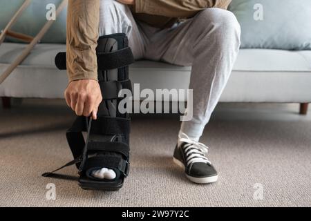 Young Man attaching an ankle orthosis to his leg to support ligaments while sitting on the sofa at home. Medicine concept. Stock Photo