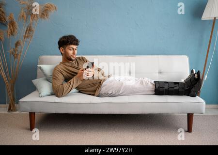 Muslim young man with ankle fracture in foot orthosis lying on sofa and using phone Stock Photo