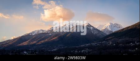 This scenic winter image features snow-capped mountain peaks surrounded by lush evergreen trees in the soft, golden light of the setting sun Stock Photo
