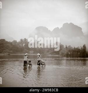 Two women wearing conical Asian hats push bicycles through a shallow river in Vang Vien, Laos. Stock Photo