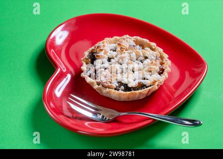 A traditional handmade mince pie and topped with flakes of marzipan served on a red heart shaped plate against a plain green background Stock Photo