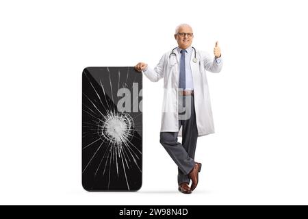 Doctor leaning on a smartphone with a cracked screen and gesturing thumbs up isolated on white background Stock Photo