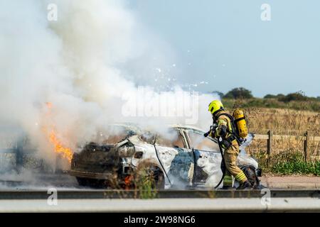 Fireman putting out a car fire on the A299, Thanet way. Spraying water into the car while smoke billows from the car and sparks fry over the top. Stock Photo