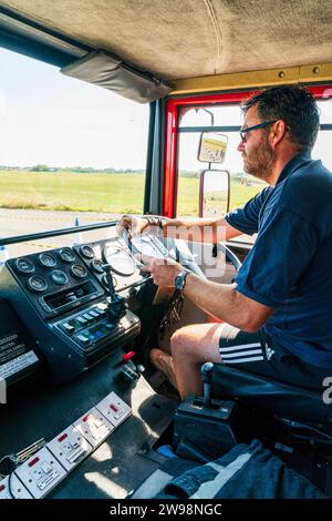 Man driving fire engine. Interior side view of man in t shirt and shorts sitting in driver's seat, both hands on the steeling wheel. Stock Photo