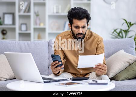 Worried young Indian man sitting on sofa at home, working on laptop, looking sadly at documents and bills, holding phone. Stock Photo