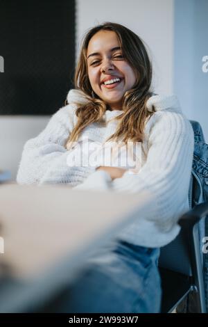 Lovely girl, junior employee, posing for the camera while sitting on a chair at the office. She is smiling. Stock Photo