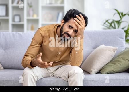 Portrait of a young Indian man looking upset at the camera, spreading his hands in frustration, suffering from a headache and depression. Stock Photo