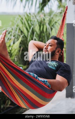 A bearded Asian man is resting in a comfortable hammock in a green garden near the paddy field. Stock Photo