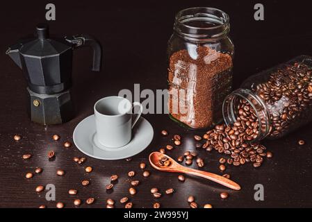 White coffee cup next to an espresso machine and two jars with ground and coffee beans, on a dark wooden table. Stock Photo