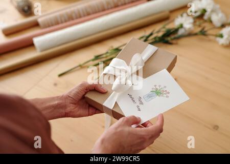 Hands of young unrecognizable woman holding handmade postcard and giftbox with white silk ribbon tied on top of cover Stock Photo
