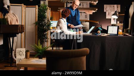 Beginner learning to design clothes, talking to experienced tailor about professional handcrafting and needlework. Senior couturier teaching apprentice the sewing process in atelier. Stock Photo
