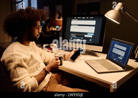 Young AI engineer with cup of coffee and smartphone in hands sitting by workplace and looking at laptop screen with data Stock Photo