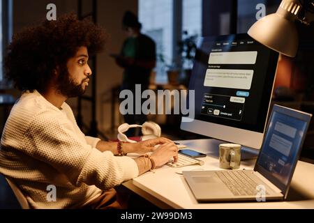 Young serious AI specialist looking at computer screen and typing on keyboard while sitting by workplace in coworking space Stock Photo