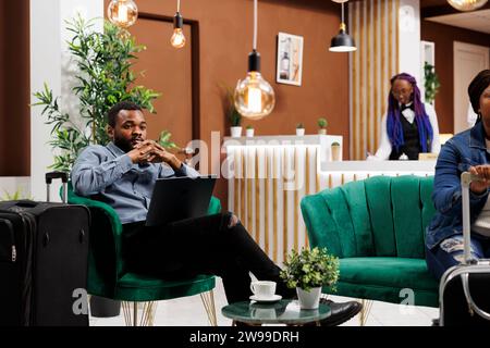 Tired African American man entrepreneur traveling on business, sitting with laptop in hotel lobby checking email. Black guy freelancer working remotely while waiting for check-in Stock Photo