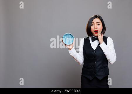 Hotel asian woman receptionist yawning while holding alarm clock and looking at camera in studio. Young attractive tired sleepy waitress oversleeping and showing time portrait Stock Photo