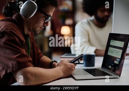 Serious young man with mobile phone texting in front of laptop with AI design tasks on screen while sitting by workplace against colleague Stock Photo