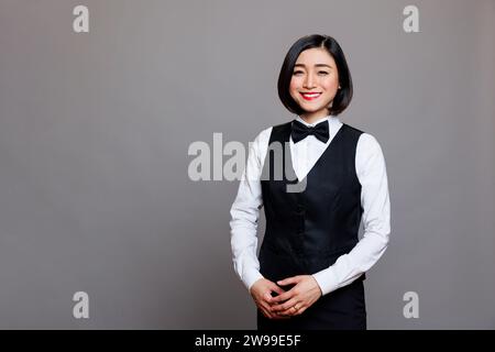 Attractive smiling asian receptionist wearing professional uniform standing and looking at camera with positive facial expression. Young waitress studio portrait on gray background Stock Photo